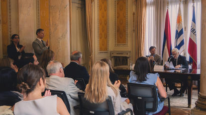 Photograph of a formal meeting in a gilded room with attendees; flags behind the speaker.