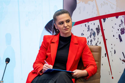 Person in a red blazer sits on a stage, holding a tablet, with an abstract mural backdrop.