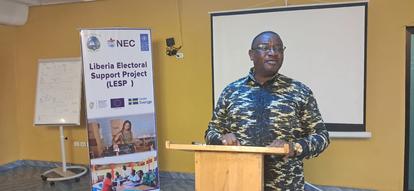 Man in a patterned shirt speaks at a wooden podium in a conference room; banner on the left.