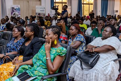 Women in colorful outfits seated in rows at an indoor event; some wearing masks.