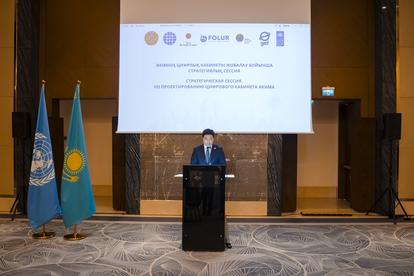 Man in a suit at podium delivering talk on stage, with flags and projection screen behind.