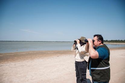Two people on a sunny beach as a photographer aims a camera toward the sea.