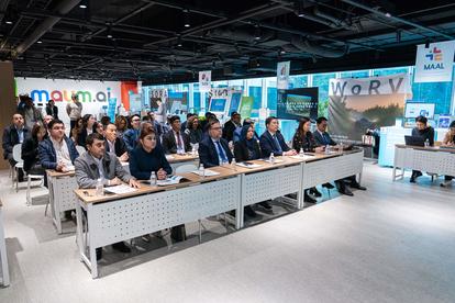Audience seated at long tables in a bright conference room with blue banners.