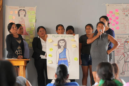 Group of students presenting colorful posters in a classroom; teacher at a podium.