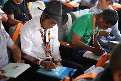 Attendee in white shirt and black hat writes in a blue notebook at a crowded conference.