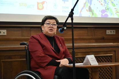 Person in a red blazer in a wheelchair on stage, speaking into a microphone, with a projection screen behind.