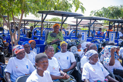 Elderly people in white sit on blue chairs outdoors; a staff member in bright shirt stands behind.
