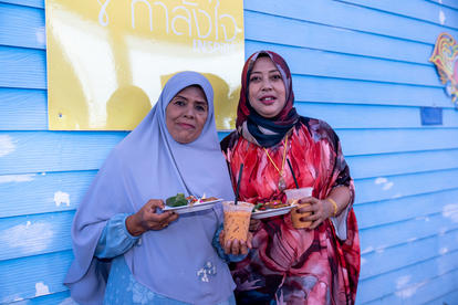 Two people in hijabs stand against a blue wall, looking at their phones.