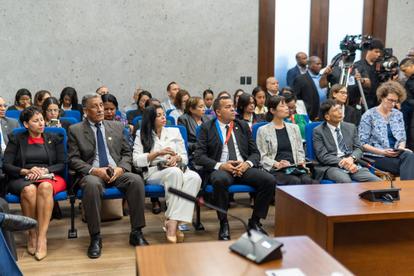 Photo shows diverse audience seated in a formal conference room, with wooden podium on the right.