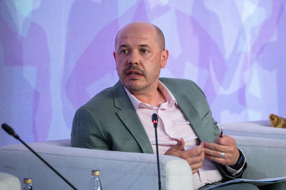 Photograph: man in a green blazer on a panel, gesturing; purple backdrop, mics