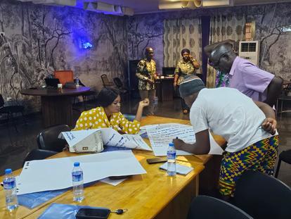 People gathered around a wooden table with laptops and papers; some wear patterned fabrics.