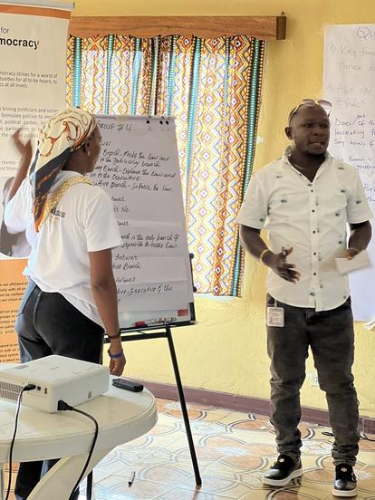 Two people presenting near a whiteboard in a sunny room with a yellow wall.