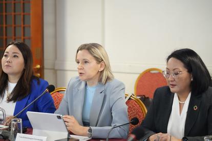 Three women in a formal meeting sit at a conference table with laptops and tablets.