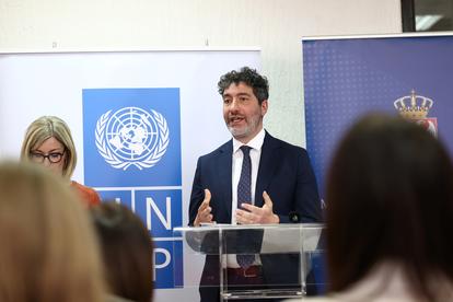Man in suit speaks at podium during UN event, blue UN banners in background.
