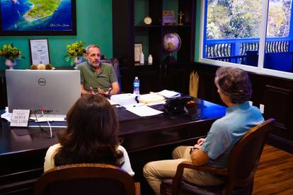 Man at a desk with a large monitor in a studio, talking to two people seated, maps on the wall.