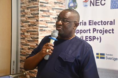 Unidentified man speaks into a microphone at an electoral support project event; brick wall and banner behind him.