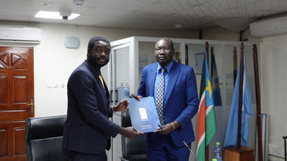 Two men in suits shake hands while presenting a document in an office with flags in the background.