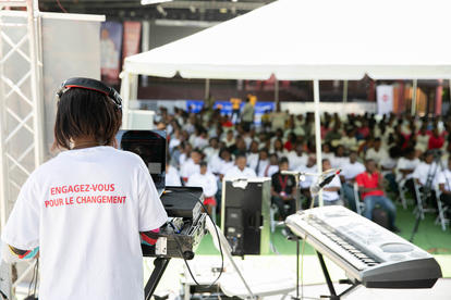 Speaker in a white T-shirt on stage with a large audience under white tents.