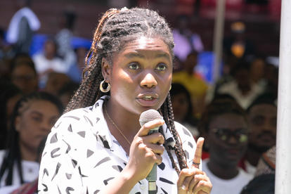 Young woman with braided hair speaks into a microphone at an outdoor event, crowd behind.