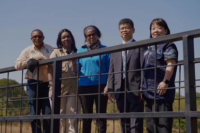 Five adults posing behind a railing outdoors, smiling, against a blue sky.