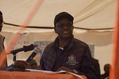 Man wearing a cap and purple jacket speaks at a table under a tent, microphones nearby.