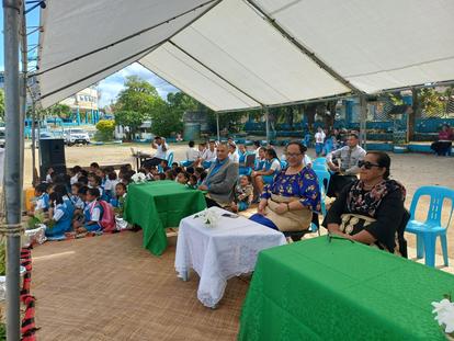 People sit under a white canopy at an outdoor community event, green tablecloths cover front tables.