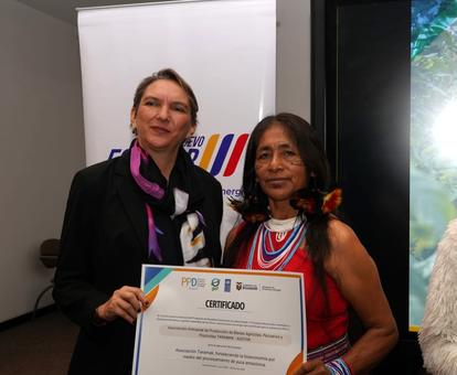 Two people hold a certificate at a formal event; faces blurred, banner backdrop.