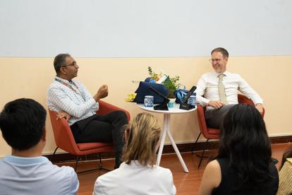 Photo: Panel discussion with two men in chairs; flowers, laptop on table; audience.