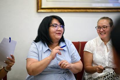Three women sit on a red couch; central woman gestures as she speaks.