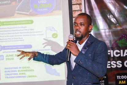 Photograph of a man in a blue suit presenting with a microphone beside a projector slide.