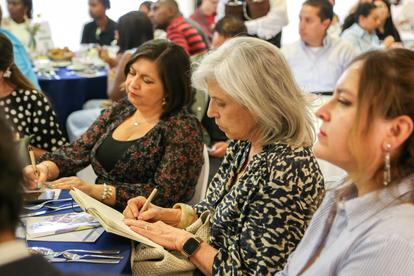 Three women sit at a blue table at a busy conference, examining papers and a smartphone.