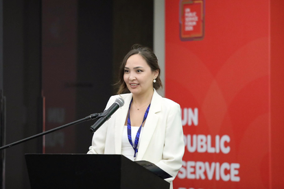 Woman in a white blazer speaks at a podium with a microphone against a red backdrop.