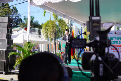 Outdoor stage under canopy; man in suit speaks, camera in foreground, banner for Entrepreneur Expo.