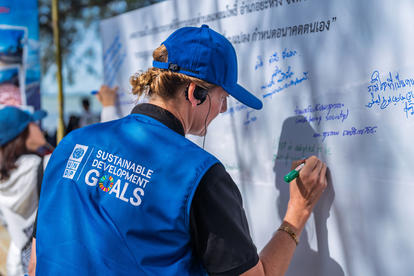 Person in a blue cap and blue vest signs a wall covered with signatures.