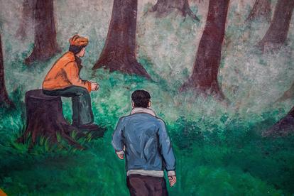Two men in a forest; one sits on a stump, the other stands with his back turned.