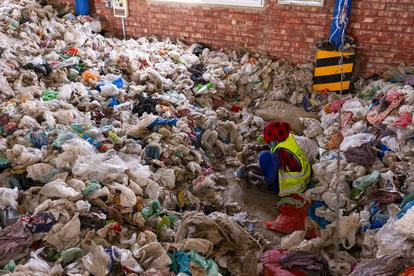Worker in a high-visibility vest sorting trash among a huge heap of garbage bags.