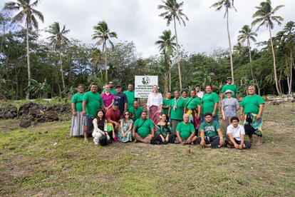Group of people in green shirts posing outdoors on a grassy field with palm trees and a sign.