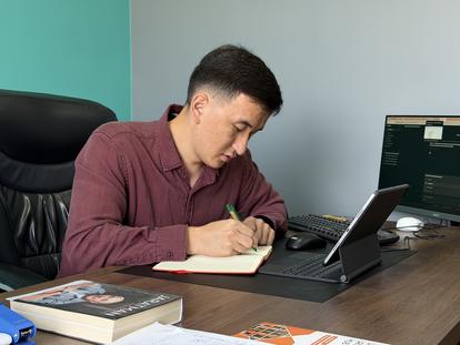 Man in maroon shirt writes in notebook at a desk with a laptop.