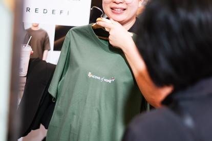 Green T-shirt with white logo worn in a clothing store; foreground shows shopper from behind.