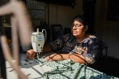 Woman wearing patterned blouse and glasses sits at a table, sewing with a vintage sewing machine.