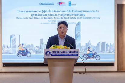 Photograph of a man in a suit giving a speech at a podium on a stage, with a screen displaying Thai text