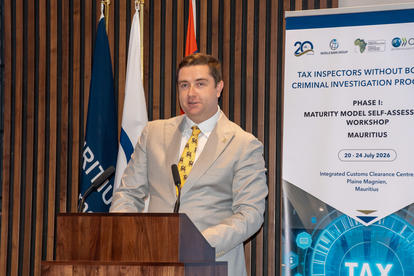 Man in a light suit and yellow tie speaks at a podium; flags and a conference banner behind.