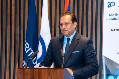 Man in suit and tie at podium delivering a speech with blue and white flags behind.