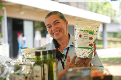 Smiling vendor at an outdoor market holds a beverage carton, jars of juice in foreground.
