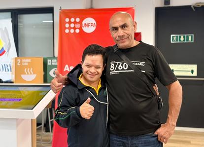 Two men pose in a store; a boy on the left gives a thumbs up to a tall man in a black shirt.