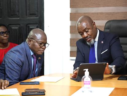 Two suited men lean over documents at a conference table with a laptop and sanitizer.