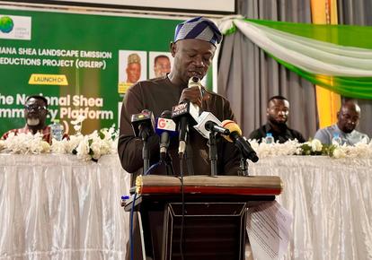 Speaker at podium with microphones during conference; green banner backdrop and panelists behind.