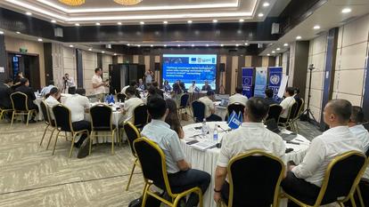 Conference room with attendees seated at tables, facing a presentation screen and banners.