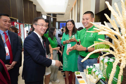 Business exhibition: man in suit shakes hands with man in green polo; woman in green smiles beside them.