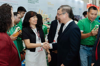 Photograph of a man in a dark suit shaking hands with a woman in white at a crowded event.